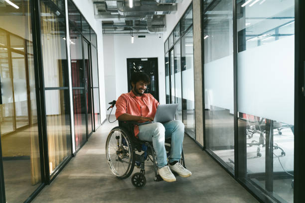 istock Disabled mixed race indian office worker in modern office corridor using laptop. Businessman in wheelchair 1355087349