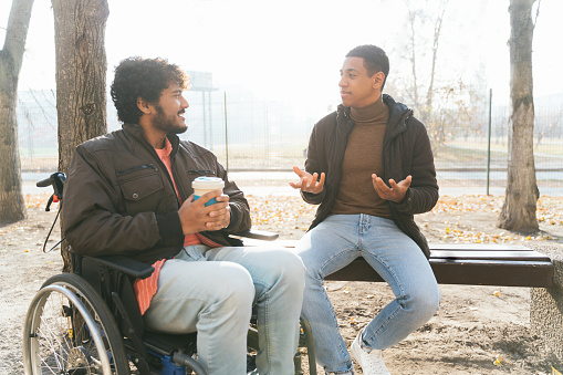 Disabled indian man in wheelchair taking walk with Latin American friend outdoors. A carer assisting disabled mixed race man