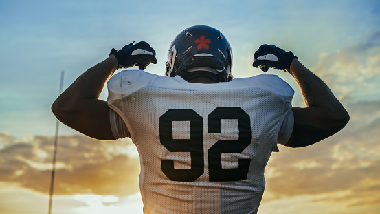 American Football Championship Game: Powerful Player Wearing Helmet, Flexing His Muscles Dramatically. Professional Athlete Determined to Win. Cheerful Sportsman Celebrating. Back View Shot