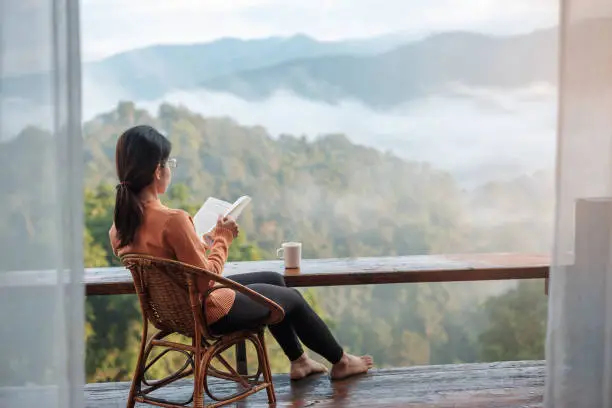 Photo of young woman reading book near window and looking mountain view at countryside homestay in the morning sunrise. SoloTravel, journey, trip and relaxing concept