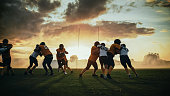 American Football Field Two Teams Compete: Players Pass and Run Attacking to Score Touchdown Points. Professional Athletes Fight for the Ball, Tackle. Dramatic Golden Hour