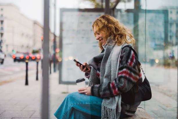 mujer joven usando un teléfono inteligente en la estación de autobuses - transporte público fotografías e imágenes de stock