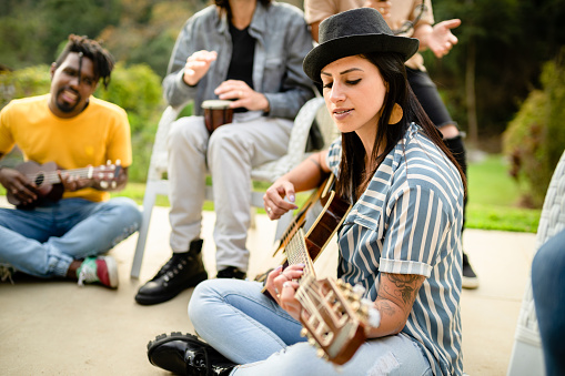 Young woman playing an acoustic guitar while jamming with a diverse group of musicians outside in summer