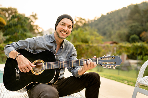 Portrait of a smiling young male musician playing an acoustic guitar outside on a patio in summer
