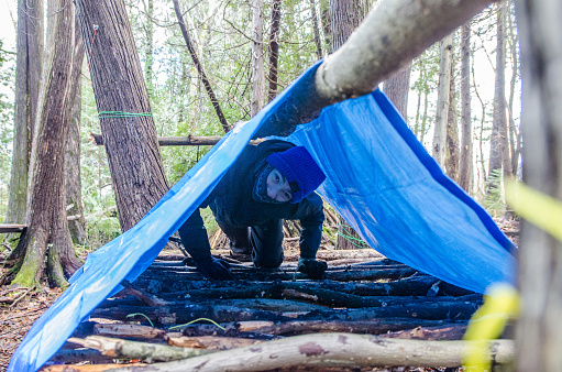 Boy building a shelter in the woods using fallen trees, rope and hand saw.
He is entering his elevated bed to sleep
He is applying survival skills.