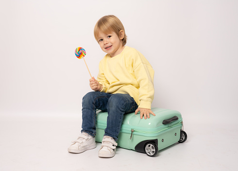 Happy little boy holding lollipop sits in a suitcase on a white background with space for text