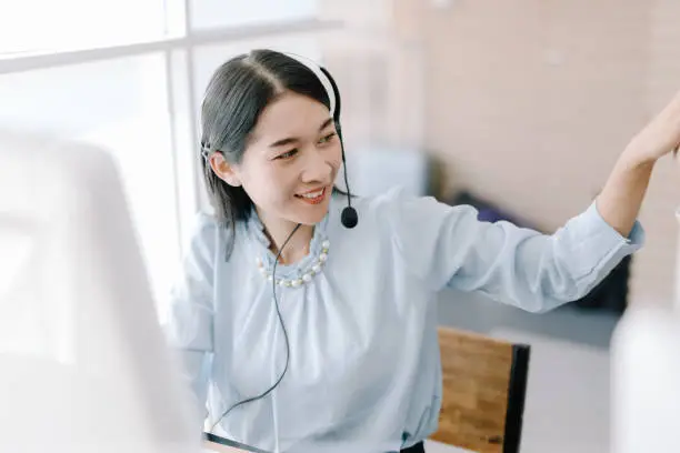 Photo of secretary operator wears a headset and microphone for consultant assistance support, the call center staff providing advice and assistance customers in smiling and happy manner while looking at camera