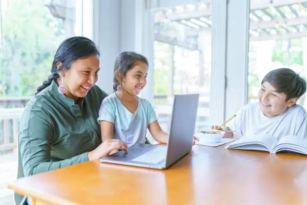 A beautiful Native American mother helps her young daughter with a school assignment online while she studies at home during the COVID-19 pandemic. The woman's adolescent son is sitting at the table working on a school assignment.