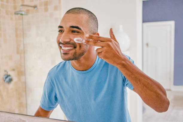 shot of a young man applying moisturizer to his face in the bathroom at home - applying imagens e fotografias de stock