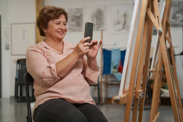 Positive delighted female person taking photo on telephone Cheerful senior woman keeping smile on her face while shooting easel with her picture painted image paintings oil paint senior women stock pictures, royalty-free photos & images