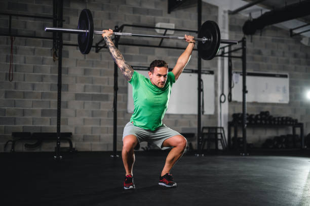 Strong Young Man Doing Barbell Overhead Squat Full length front view of Caucasian man in late 20s squatting while holding barbell and weights overhead in gym. deadlift alternatives stock pictures, royalty-free photos & images