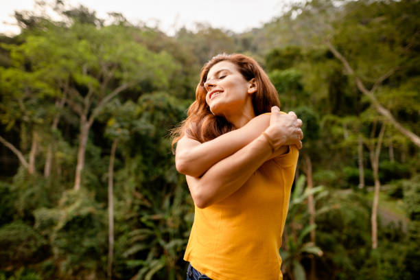 Smiling woman giving herself a hug outside in summer Smiling woman standing with her eyes closed and giving herself a self-love hug outside by a forest older woman eyes closed stock pictures, royalty-free photos & images