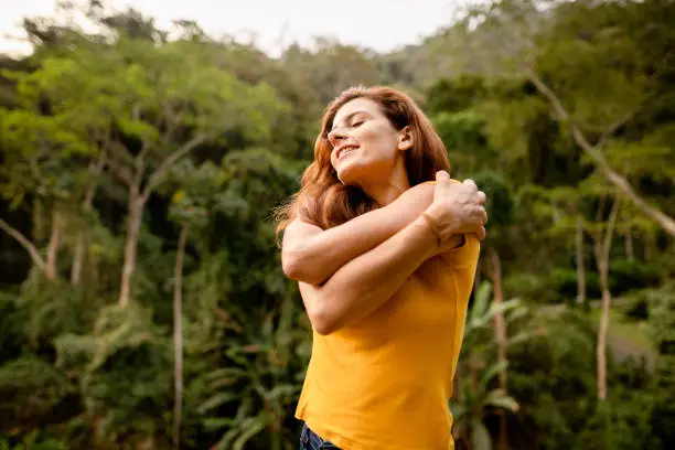 Smiling woman standing with her eyes closed and giving herself a self-love hug outside by a forest