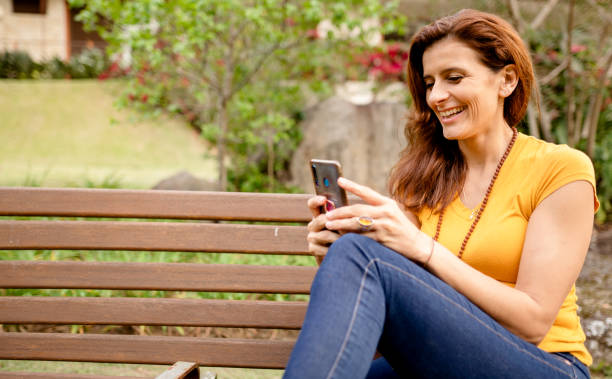 smiling woman sitting on a bench in her yard and texting on her phone - using phone garden bench imagens e fotografias de stock