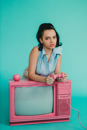 Portrait of a beautiful young retro-styled woman gaming on a pink color vintage TV and pink color gamepad, studio shot in front of a blue background