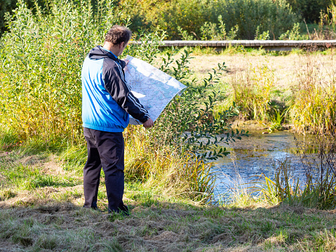 Man standing by river and holding map, middle age tourist enjoying sunny day in autumn nature and looking in map