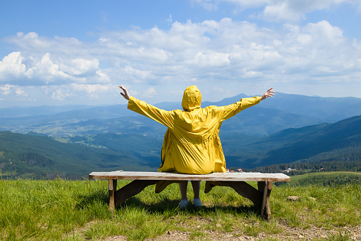 Adventure female hiker raising hands sits on a bench and enjoying mountain panorama view on a sunny summer day