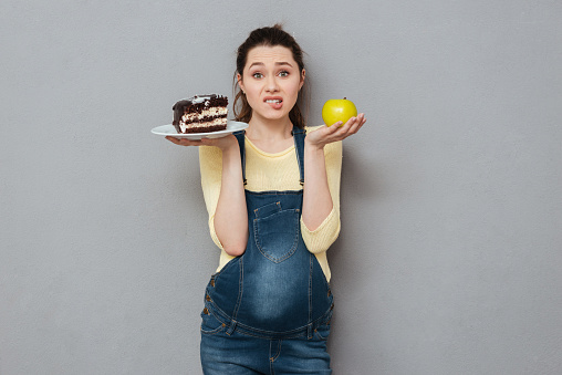 Image of pregnant lady standing isolated over grey wall while choosing between sweet cake and apple. Looking at camera.