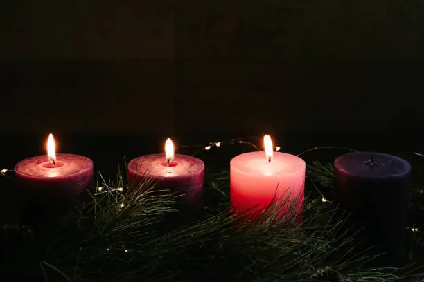 Three pillar candles lit in an advent wreath, two purple and one pink, for the third week of advent, gaudete Sunday. With evergreen boughs and copy space in the dark.