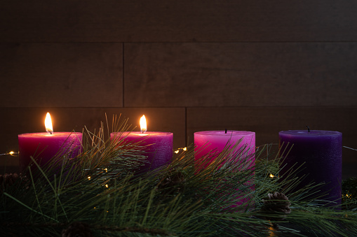 Two advent pillar candles burning for the second week of advent, in a dark room with copy space against a wood background with evergreen branches of an advent wreath.