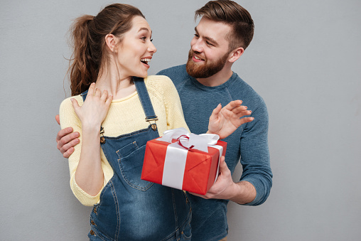 Portrait of a smiling husband giving gift box to his pregnant wife isolated over gray background