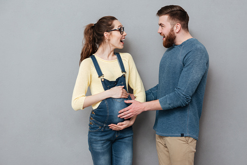 Image of pregnant cheerful woman wearing glasses standing near happy man isolated over grey wall.