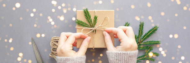Banner with Female hands decorate a Christmas gift box, string and spruce branch on a gray background. Ecological concept. Flat lay, copy space.