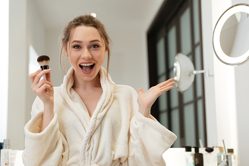 Happy amazed young woman applying powder brush and doing makeup in bathroom