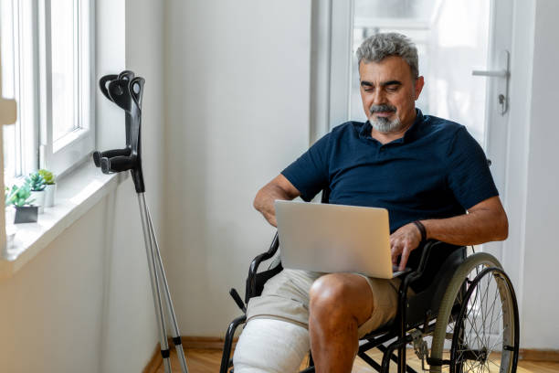 senior man with broken leg on wheelchair - laptop mature adult senior adult old imagens e fotografias de stock