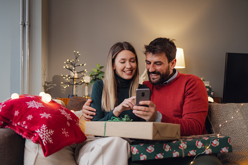 Couple sitting in the living room and celebrating Christmas holidays and holding gifts while using mobile phone