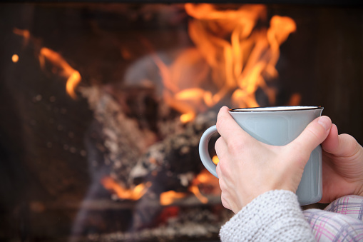 Close-up of woman hands holding tea cup in front of fireplace at home