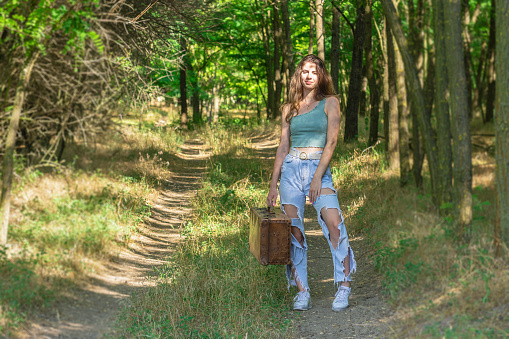 Young woman standing in forest dirt road and holding an old brown suitcase