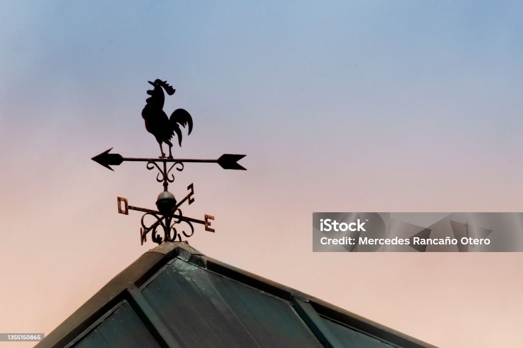 Weathervane in rooster shape, sunset background. Close up of weathervane in rooster shape silhouetted on a rooftop, sunset sky in the background with copy space on the right.  Tui, Pontevedra province, Galicia, Spain. Weather Vane Stock Photo