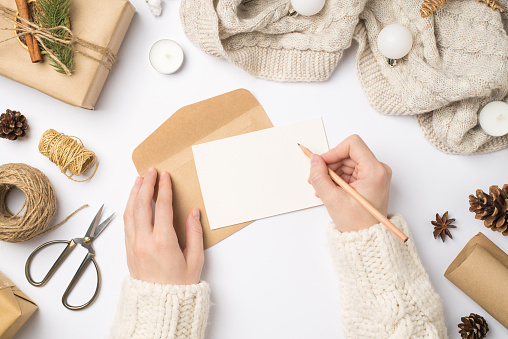 First person top view photo of hands in sweater writing letter giftbox scarf christmas tree balls handicraft tools cones anise candles scissors spool of twine isolated white background with copyspace