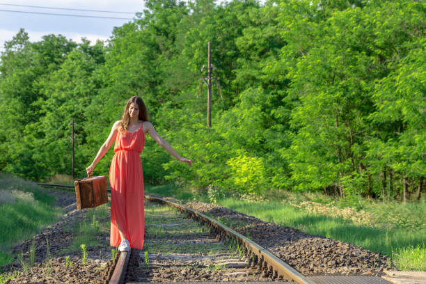 Young woman in red dress balancing on rails with old brown suitcase