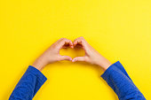 Kid hands making a heart shape on yellow background. Top view