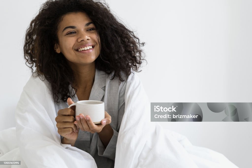 African american woman sitting with cup of coffee Happy and joyful african american woman wrapped in soft blanket, sitting in pajamas, holding cup of coffee in hands, smiling wide against white copy space background. Recreation, relaxation concept Blanket Stock Photo