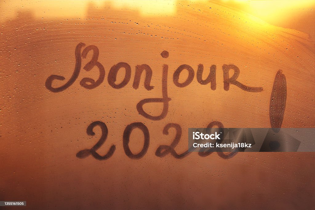 lettering Bonjour in French is hello in english and numbers 2022 paint with finger with streaks of water on splashed by rain foggy glass on orange sunset window Handwritten lettering Bonjour in French is hello in english and arabic numerals 2022 on misted glass on orange sunset window flooded with raindrops 2022 Stock Photo