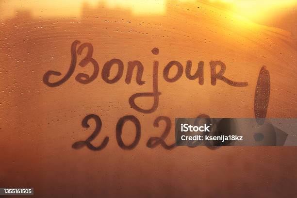 Lettering Bonjour In French Is Hello In English And Numbers 2022 Paint With Finger With Streaks Of Water On Splashed By Rain Foggy Glass On Orange Sunset Window Stock Photo - Download Image Now