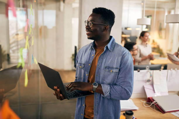 istock Happy black entrepreneur using laptop while examining mind map on glass wall at casual office. 1355163625