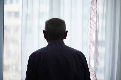 Silhouette of retired man looking through window with transparent curtain