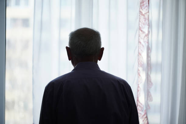 Silhouette of retired man looking through window with transparent curtain Silhouette of retired man looking through window with transparent curtain standing at home rear view. Loneliness and old human care concept. loneliness stock pictures, royalty-free photos & images