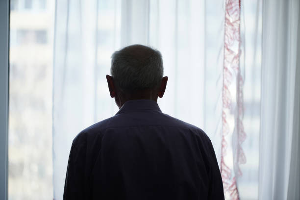 Silhouette of retired man looking through window with transparent curtain Silhouette of retired man looking through window with transparent curtain standing at home rear view. Loneliness and old human care concept. one senior man only stock pictures, royalty-free photos & images