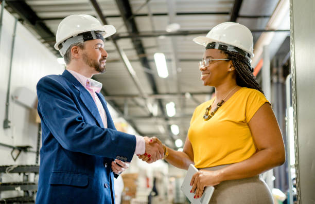 Two smiling diverse businesspeople shaking hands in a large warehouse Two diverse businesspeople wearing hardhats smiling and shaking hands together while standing in a large warehouse business relationship stock pictures, royalty-free photos & images