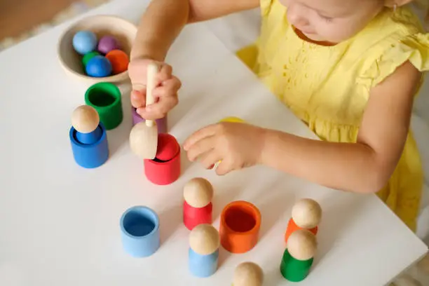 Toddler plays with different figures at the table, sorting them by color, throwing a ball with a spoon into a cup. Early development of a baby using games and toys. Logical thinking and Entertainments