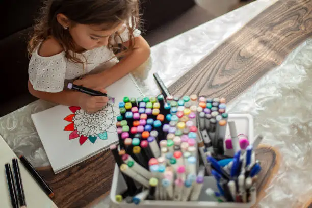 Photo of A little cute girl draws a circular mandala pattern in the album with art markers