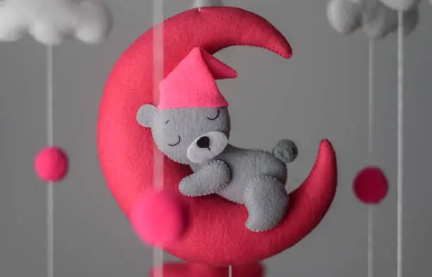 Sleeping bear on the moon, felt cot mobile close up, soft plushie toy decoration over the crib in the baby room. Pink hat on the teddy bear. Childhood development concept.