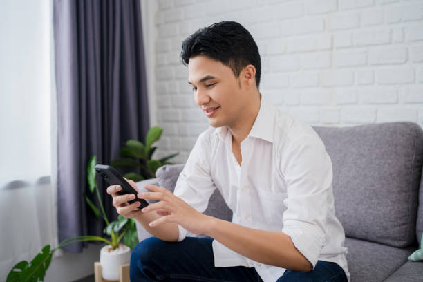 istock Happy smiling young male using smartphone while sitting and enjoying on comfortable couch at home 1355216395