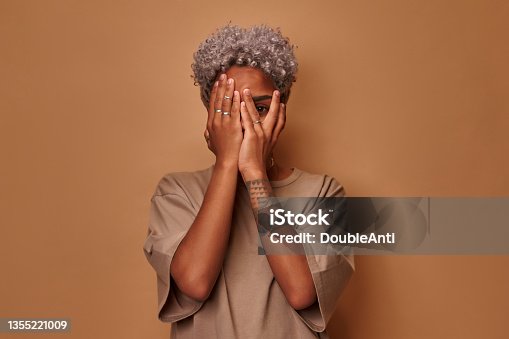 istock Frightened dark skinned woman covers face with palms peeks through fingers 1355221009