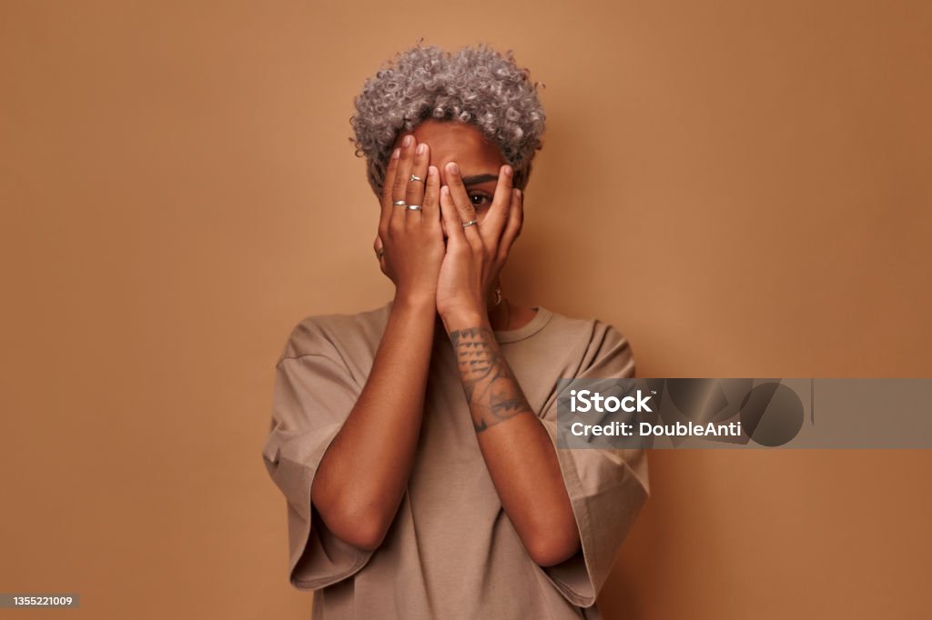 Frightened dark skinned woman covers face with palms peeks through fingers Frightened dark skinned woman covers face with palms peeks through fingers sees something horrible and unexpected, hides from all difficulties intrigued what happening, against studio beige background Fear Stock Photo